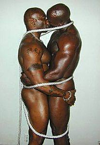 Two muscular Black men are bound together with white rope while embracing and kissing against a plain wall.