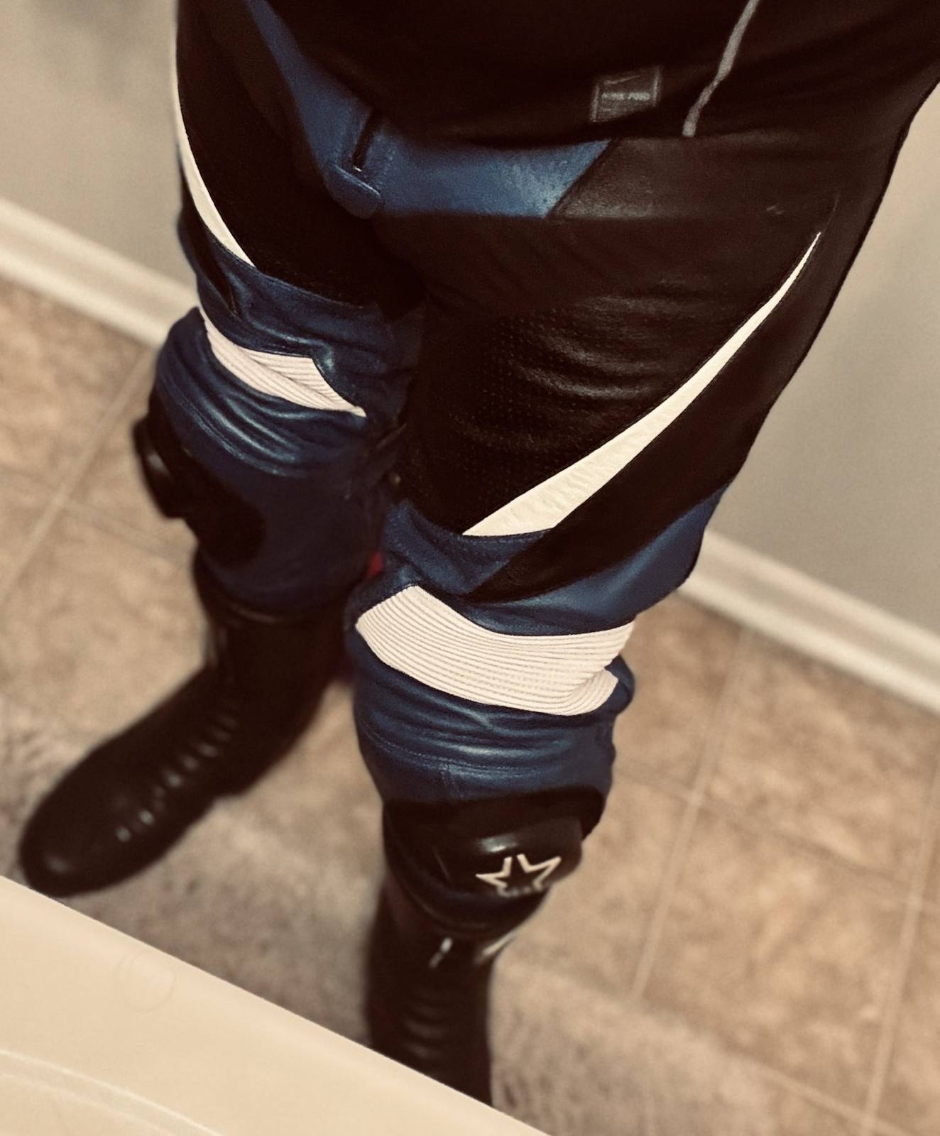 A man wearing blue, black, and white motorcycle leathers paired with tall black boots stands in a tiled room.
