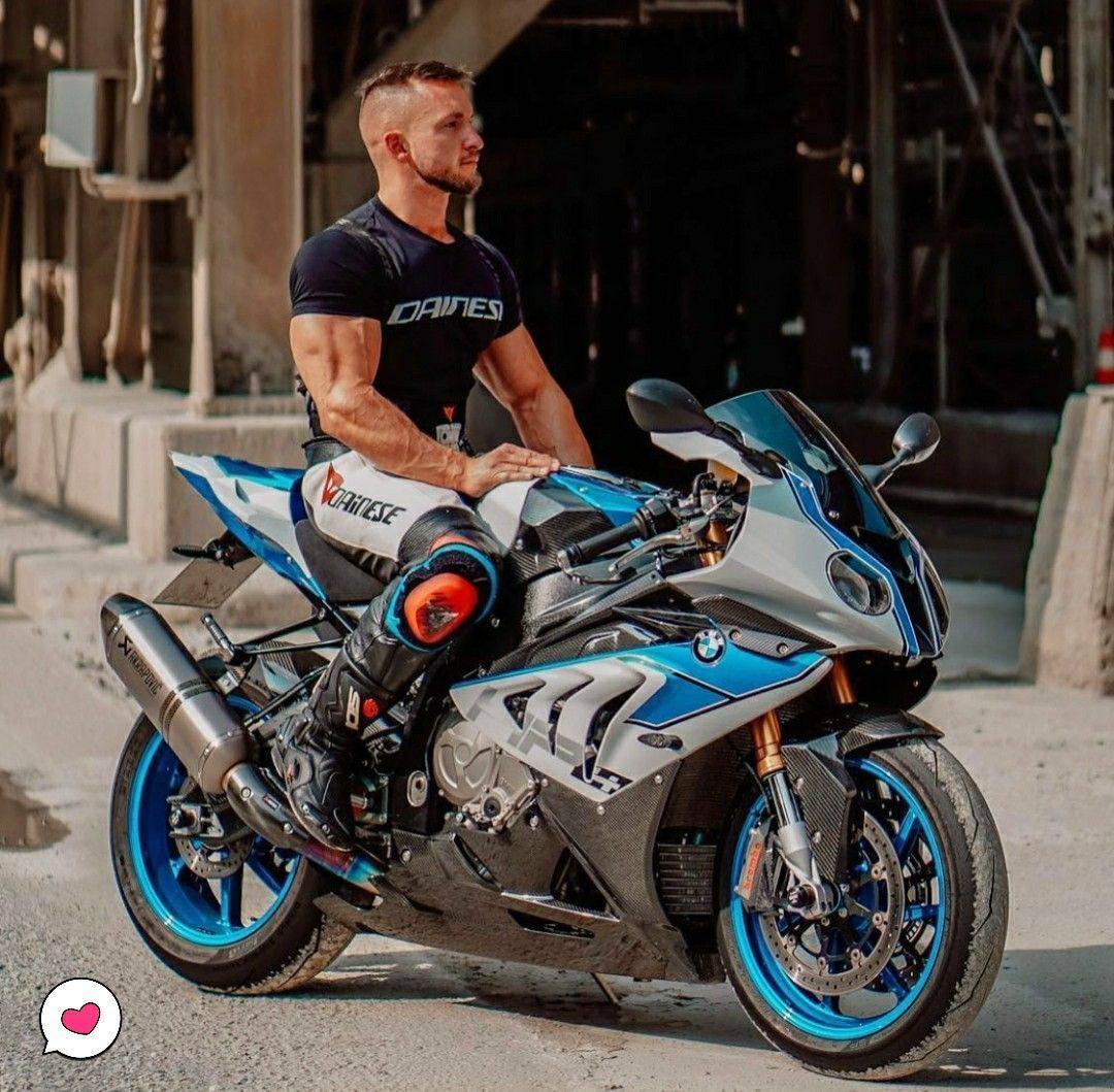 A muscular man with a beard and buzzcut poses on a sport motorcycle wearing leather racing pants and boots.