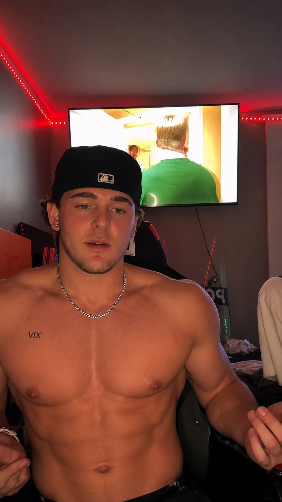 A shirtless muscle stud with a silver chain and a black baseball cap poses in a room with red ambient lighting.