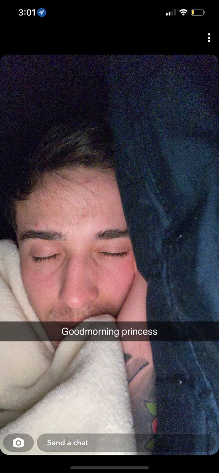 A young man with dark hair and light stubble sleeps in bed under a white blanket with a tattooed arm resting beside him.