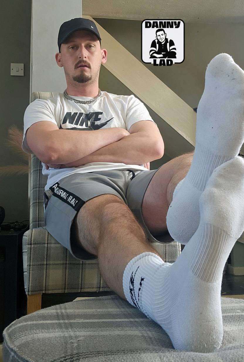 A bearded man wearing a Nike t-shirt and grey gym shorts poses with his feet in white crew socks propped up.