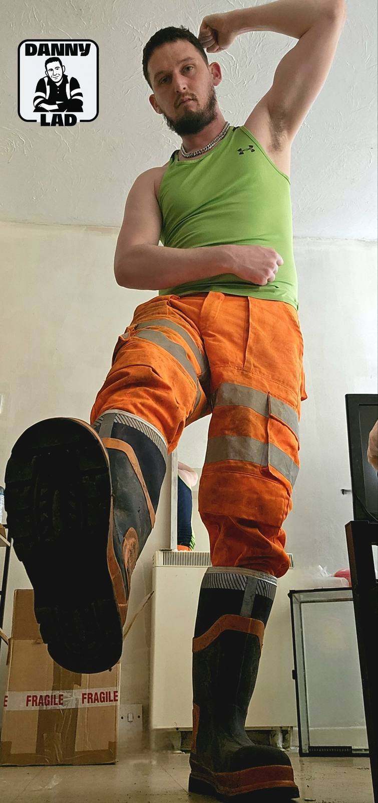 A bearded man wearing a green tank top and orange hi-vis work trousers poses with a large rubber boot raised toward the camera.