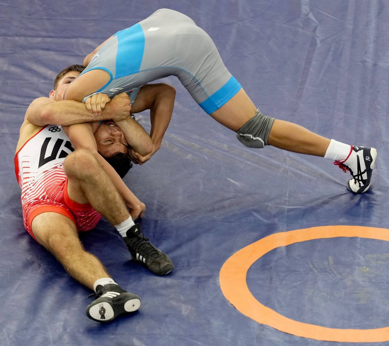 Two athletic young men in wrestling singlets are shown grappling and pinning each other on a blue competition mat.