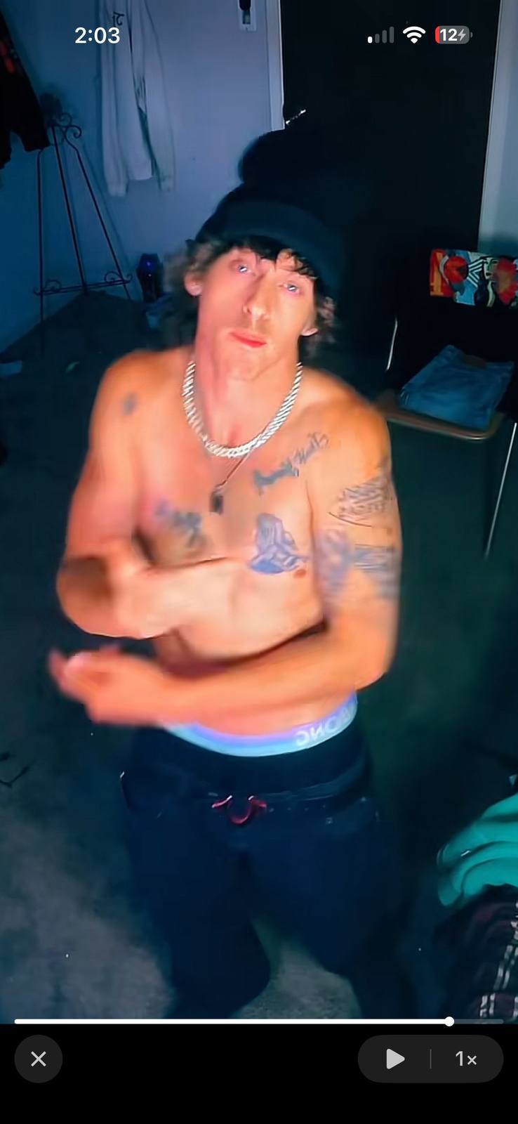 A shirtless tattooed twunk wearing a black beanie and sagging jeans poses to show his underwear waistband.