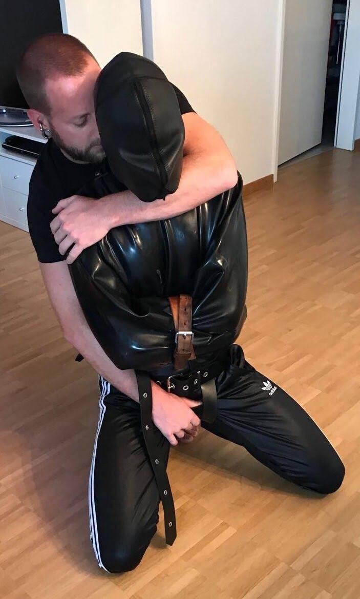 A bearded man stands behind a submissive who is kneeling in a shiny latex restraint jacket and a full leather sensory deprivation hood.