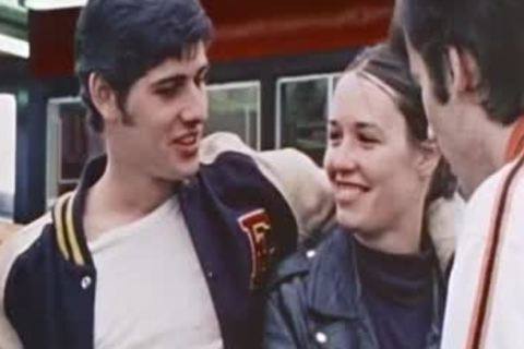 A young man wearing a classic varsity letterman jacket stands outdoors alongside friends in leather gear.