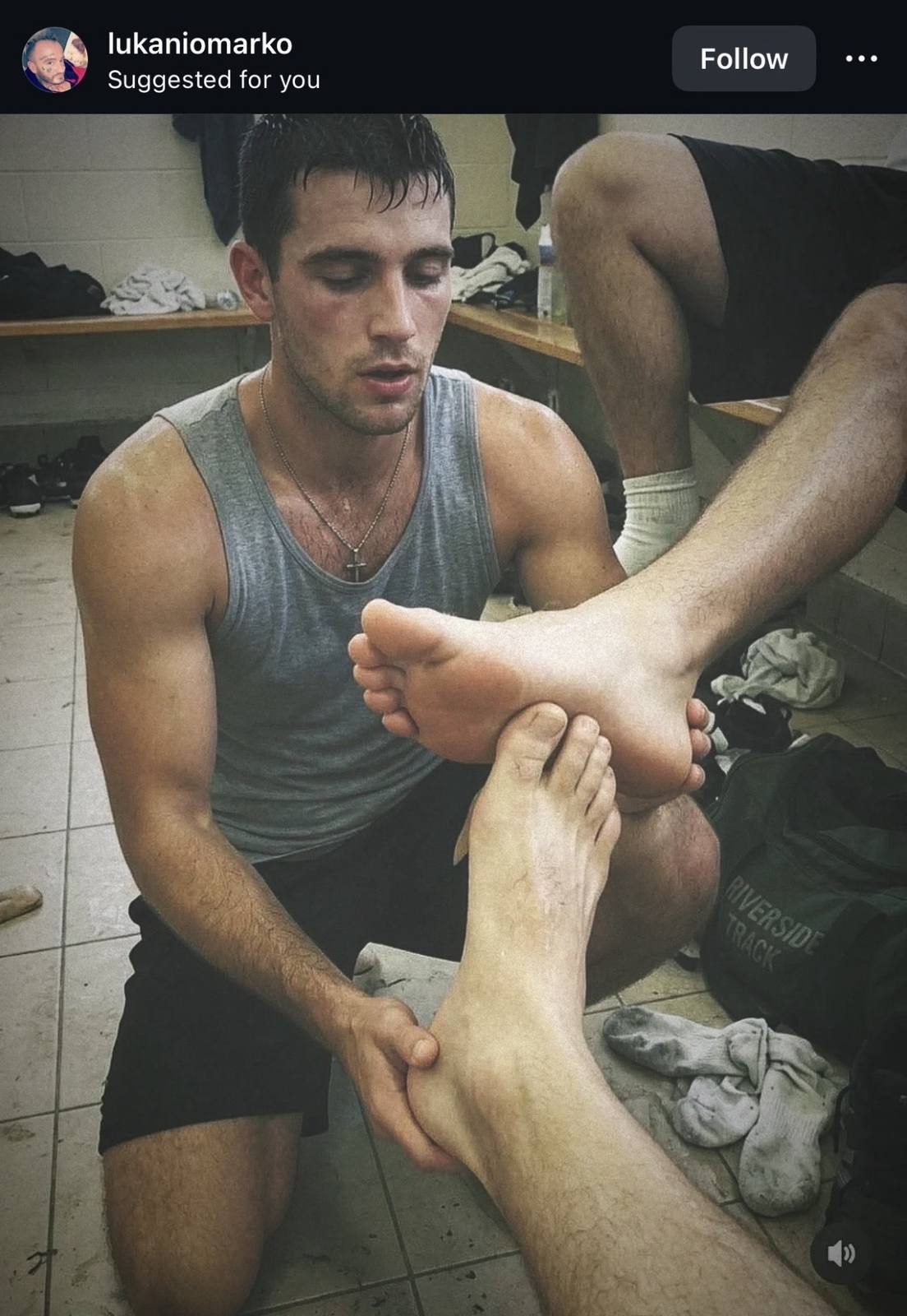 A muscular jock in a grey tank top kneels in a gym locker room to worship and massage another man's sweaty feet.