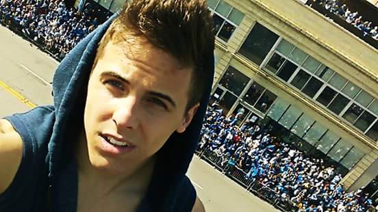 A young man takes a selfie outdoors in front of a large crowd.