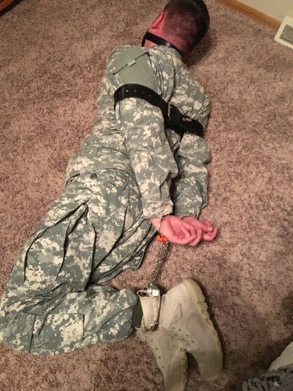 A man in a military camouflage uniform is restrained on a carpeted floor with a ball gag, handcuffs, and leather straps.