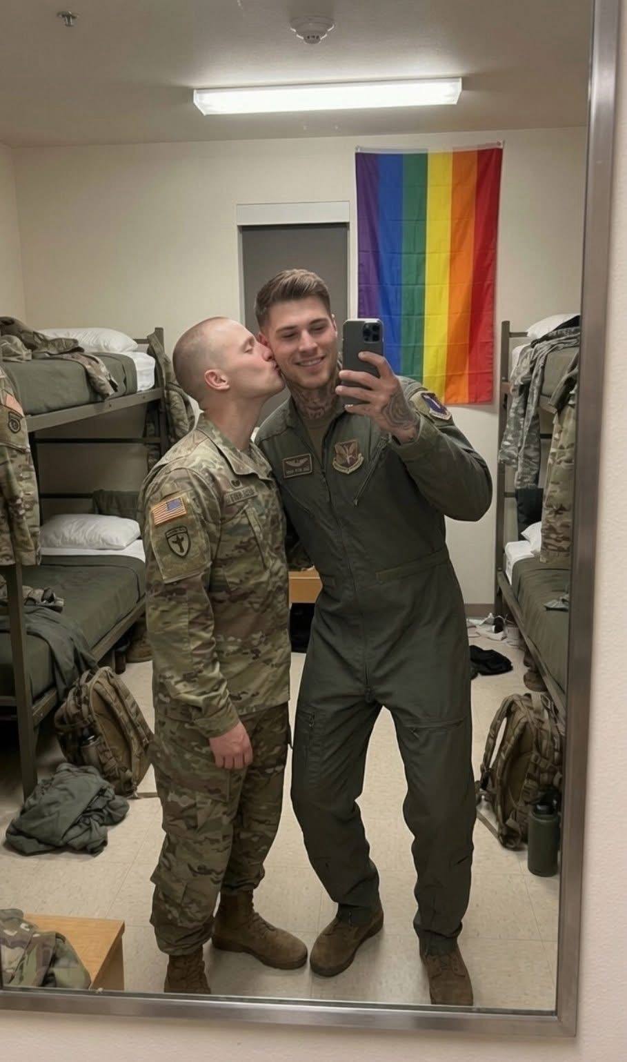 Two men in military uniforms, one in camo fatigues and the other in a green flight suit, share a kiss in a barracks room featuring a pride flag.