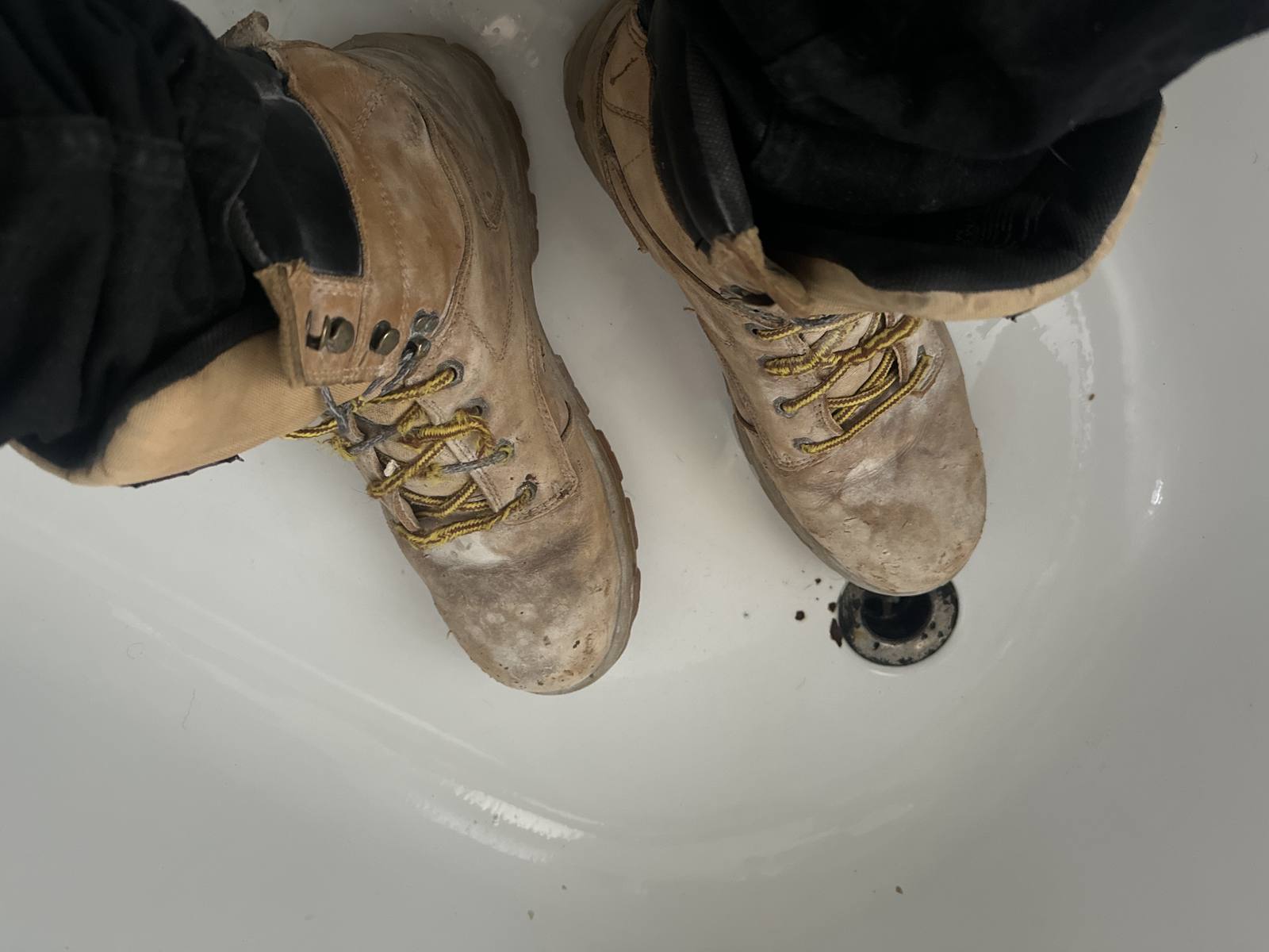 A top-down POV shot shows a person wearing scuffed and dirty tan work boots while standing in a white bathtub.