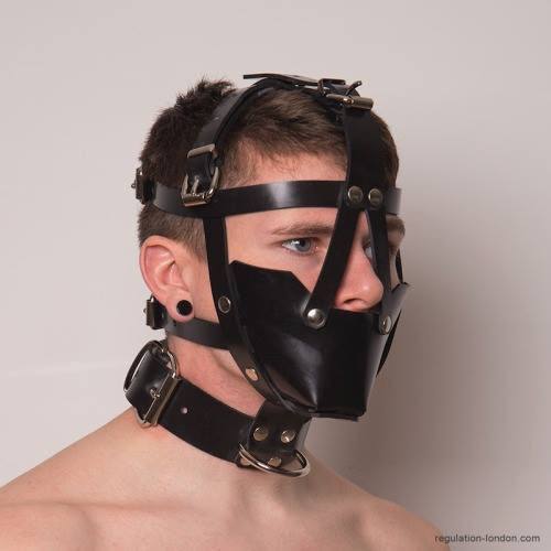 A young man wears a black leather head harness muzzle and a heavy leather collar against a plain studio backdrop.
