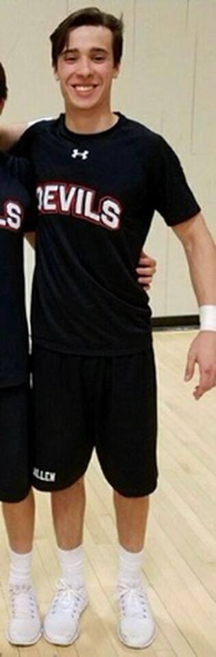 A smiling young man stands in a gymnasium wearing a black athletic t-shirt, black gym shorts, white crew socks, and white sneakers.
