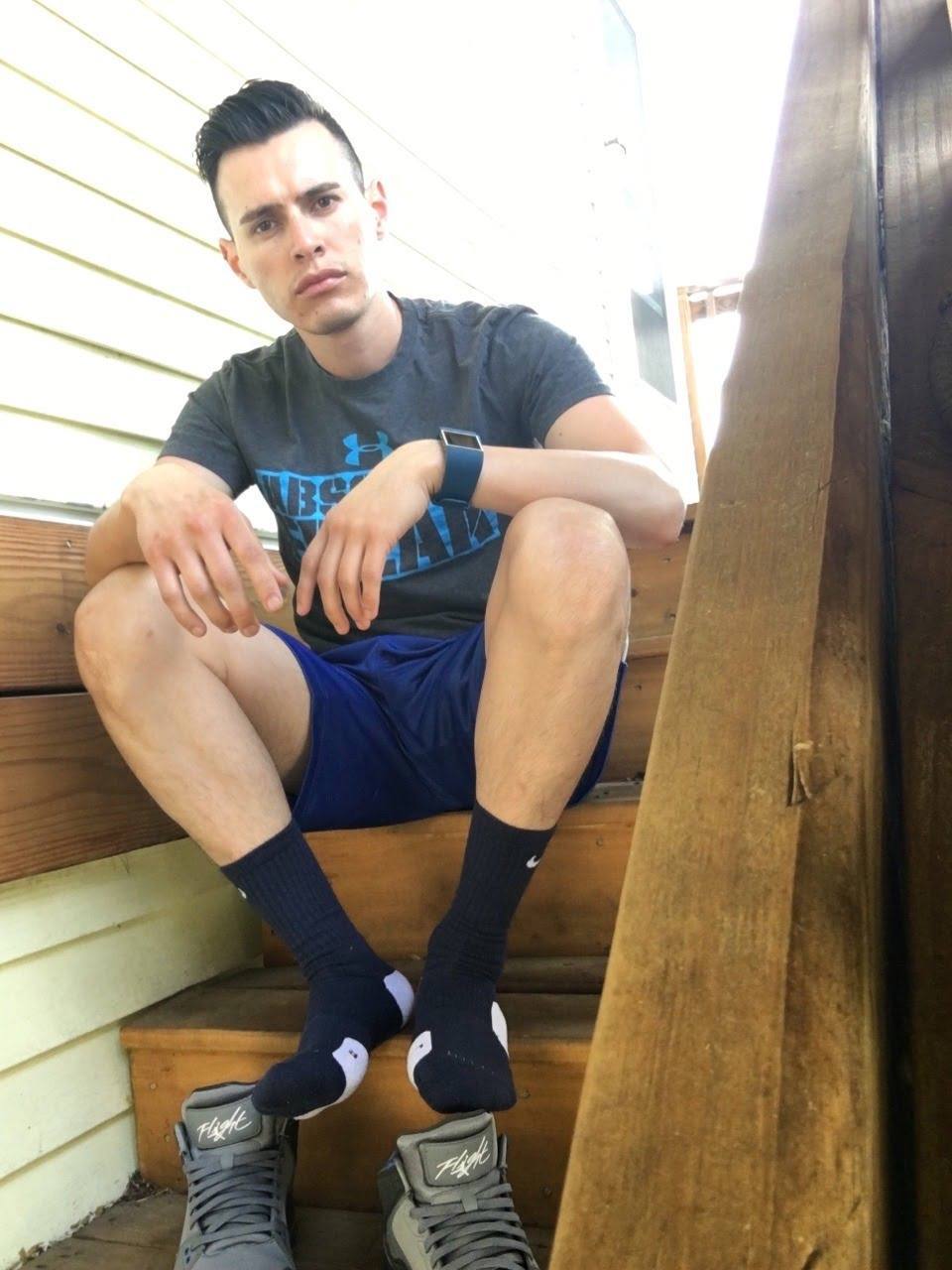 A young man sits on outdoor wooden stairs wearing a t-shirt, blue gym shorts, and dark crew socks with his high-top sneakers placed beneath his feet.