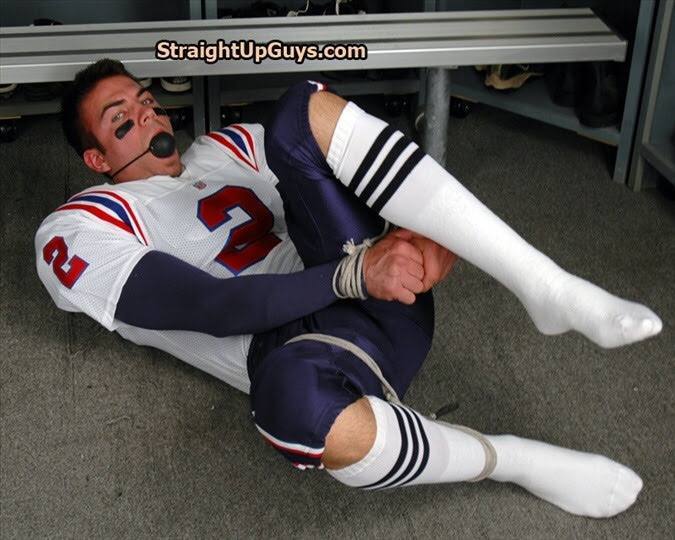 A man dressed in a football uniform lies bound with rope and gagged with a ball gag on a locker room floor.