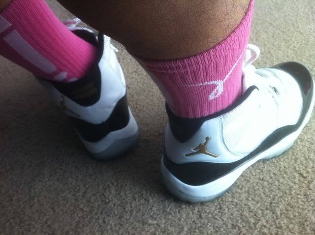 A close-up view of hairy legs wearing bright pink crew socks and white and black Jordan 11 sneakers on a carpeted floor.