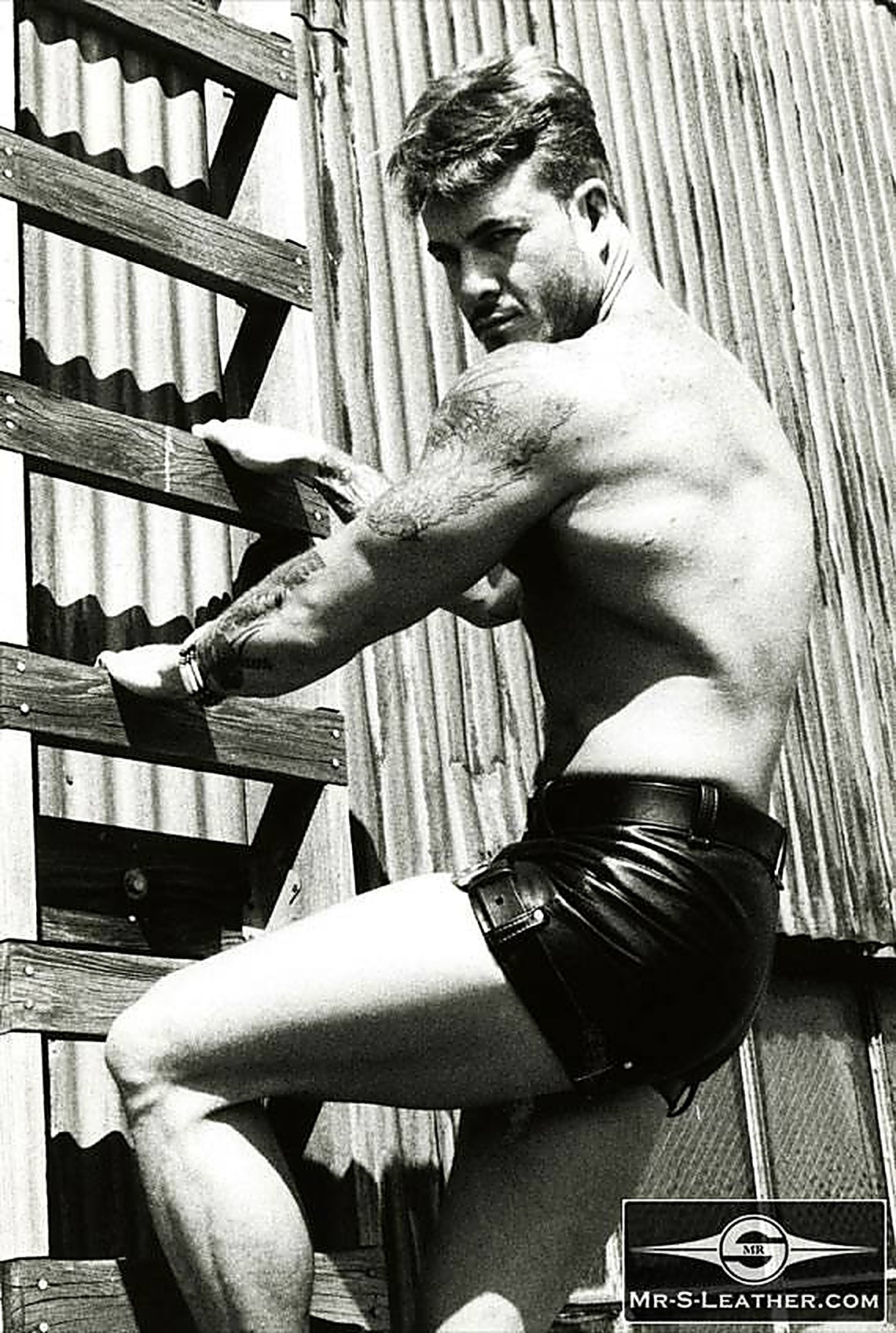 A muscular tattooed man wearing tight black leather shorts climbs a wooden ladder against a corrugated metal wall.