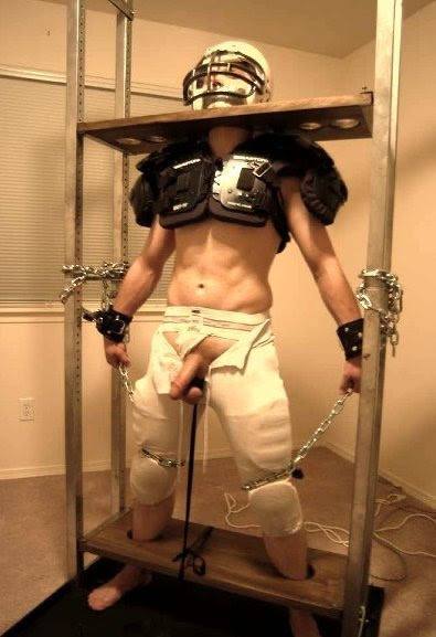A toned man wearing football shoulder pads and a helmet stands locked in a tall wooden pillory frame with exposed genitals.