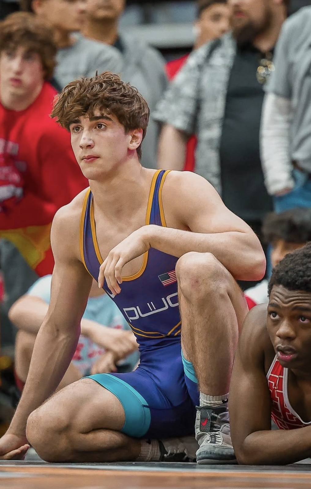 A muscular young wrestler wearing a tight purple spandex singlet kneels on the mat during a match.