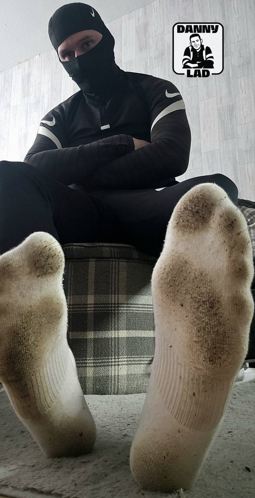 A man wearing black athletic gear and a balaclava sits with his legs open, showing the soles of his dirty white socks to the camera.