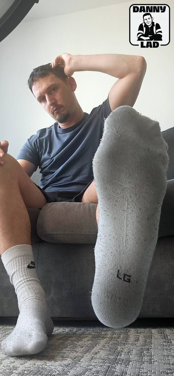 A man in a t-shirt and gym shorts sits on a sofa, extending a white crew sock-covered foot towards the camera in a POV perspective.