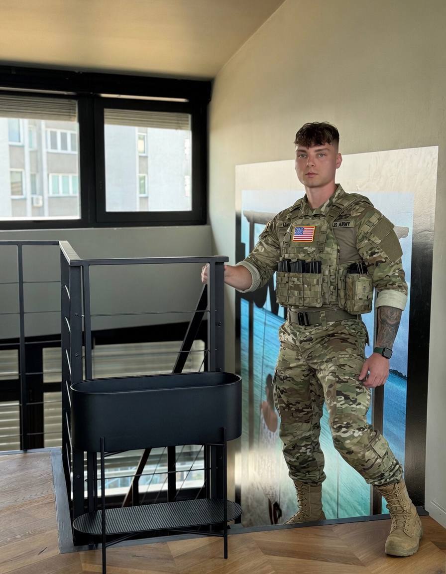 A fit young man with a buzzcut and arm tattoos poses indoors wearing a full military camouflage uniform and tactical vest.