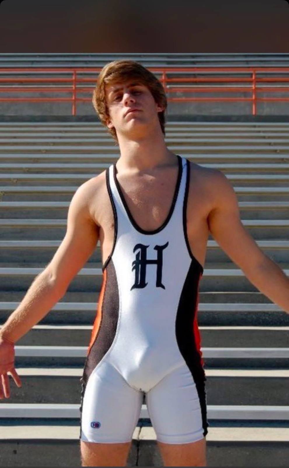 A young, athletic man poses in a tight white and black wrestling singlet in front of stadium bleachers.