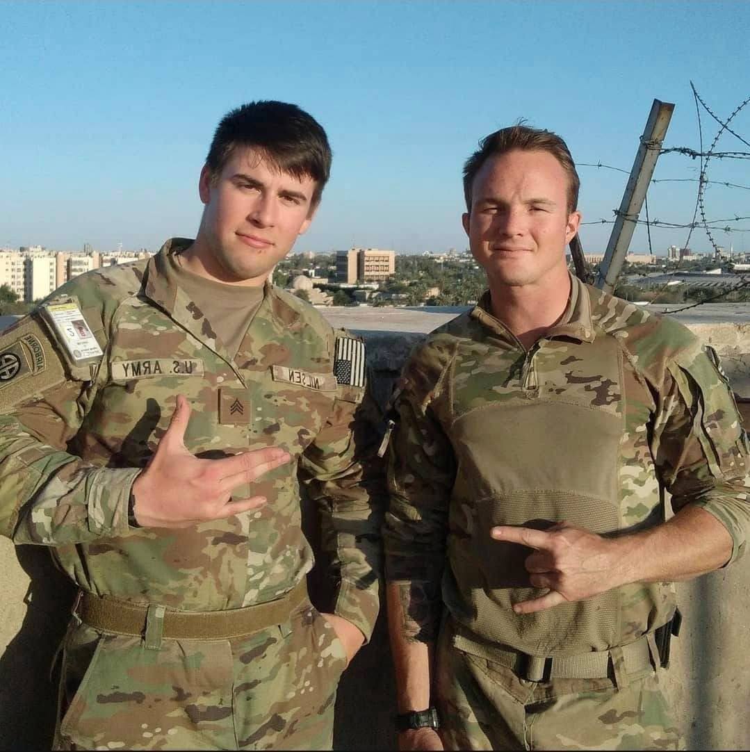Two fit men in US Army camouflage uniforms pose together on an outdoor rooftop overlooking a city.