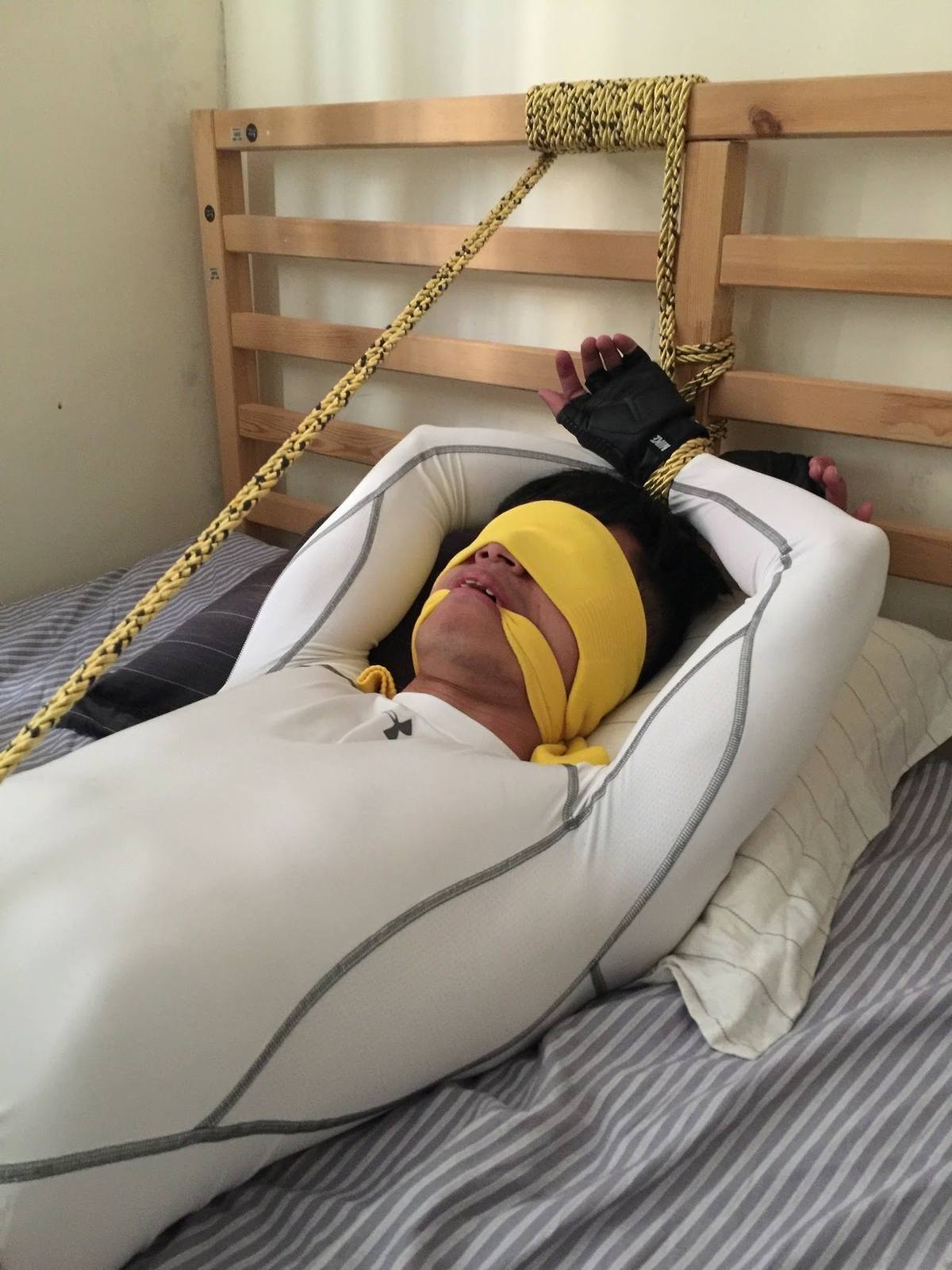 A young man wearing a white compression shirt and fingerless gloves lies bound to a wooden headboard with yellow rope, blindfolded and gagged.