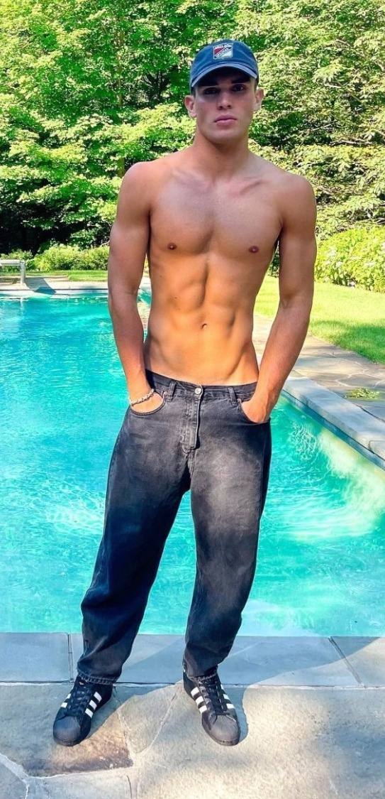 A muscular, shirtless young man poses in black jeans and a baseball cap beside an outdoor swimming pool.