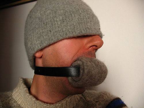A man is gagged with a rolled grey wool sock secured by a black leather strap while wearing a grey beanie pulled down over his eyes.