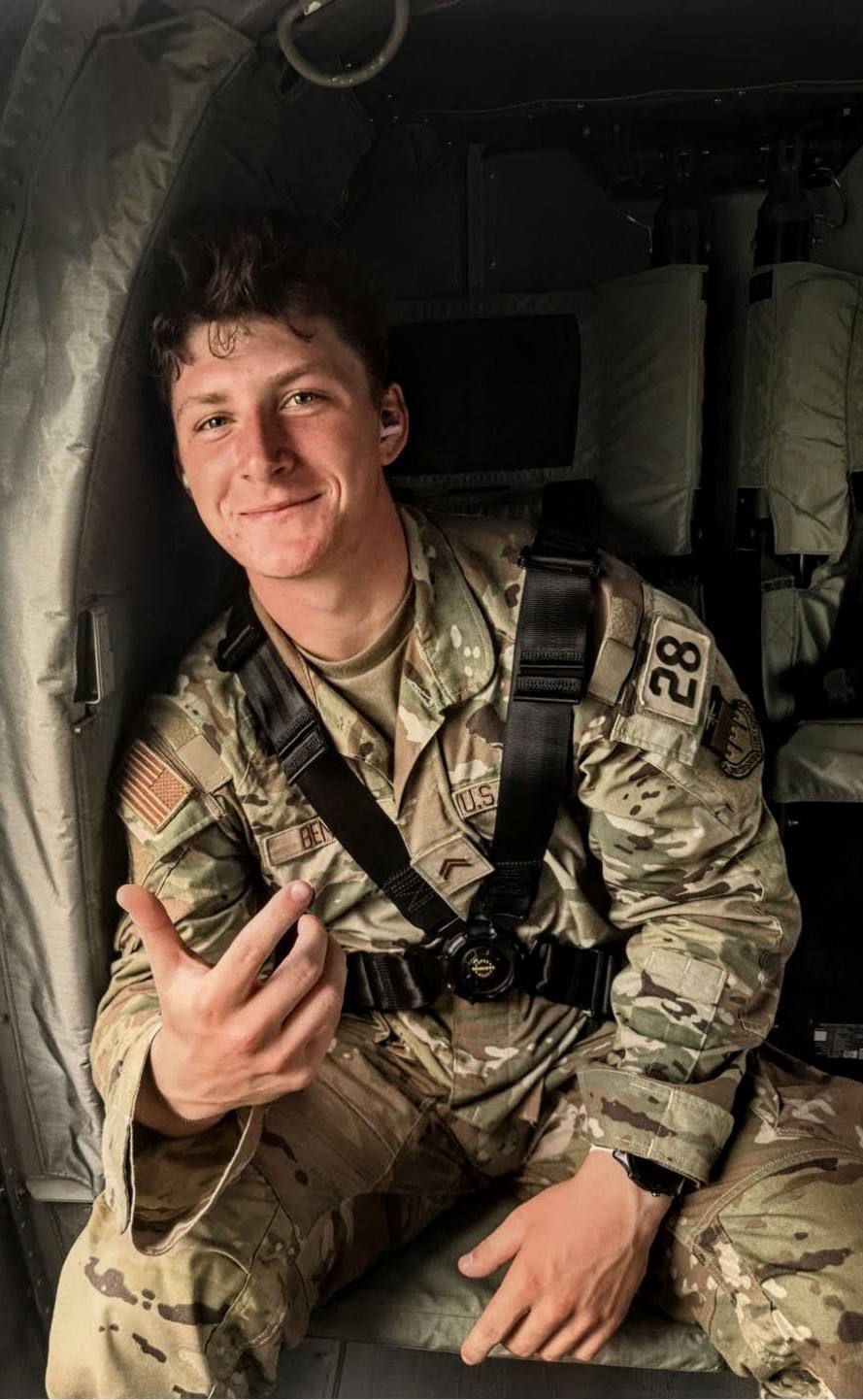 A young man in a camouflage military uniform and black safety harness sits inside a military aircraft.