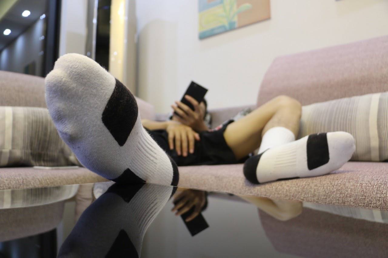 A person wearing white athletic socks and black gym shorts relaxes on a sofa while looking at a phone.