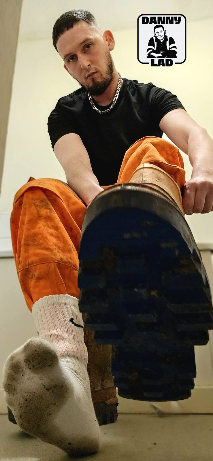A man in orange work trousers and a black t-shirt presents the sole of his heavy work boot and a dirty white tube sock toward the camera.