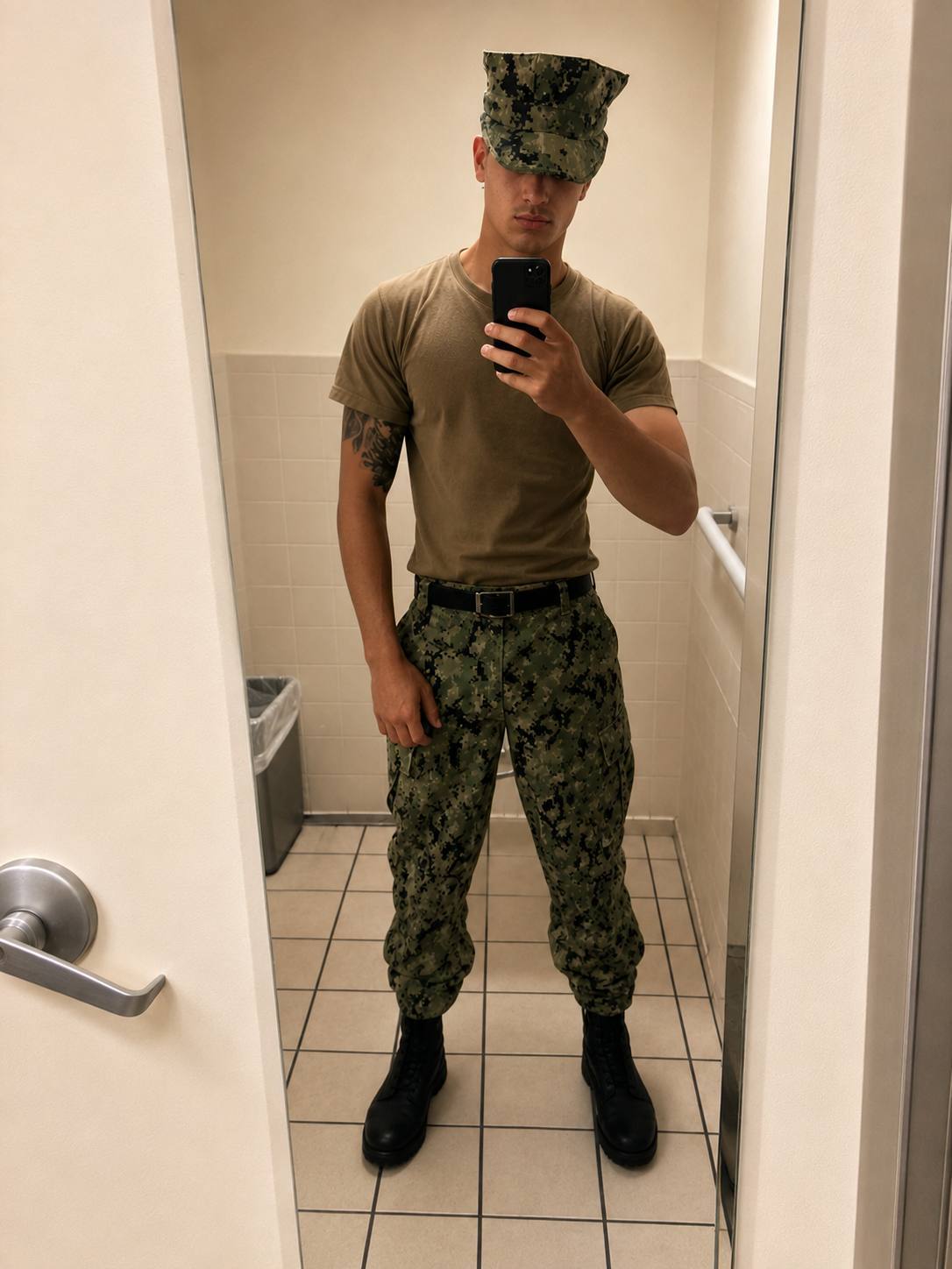 A young, athletic man in a green camouflage military uniform and combat boots takes a mirror selfie in a restroom.