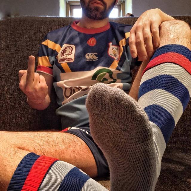 A bearded man wearing a Queensland Rugby jersey and striped socks extends one foot toward the viewer while making a gesture.