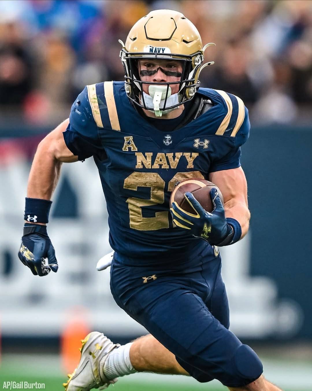 A muscular American football player in a Navy uniform runs with the ball on the field during a game.