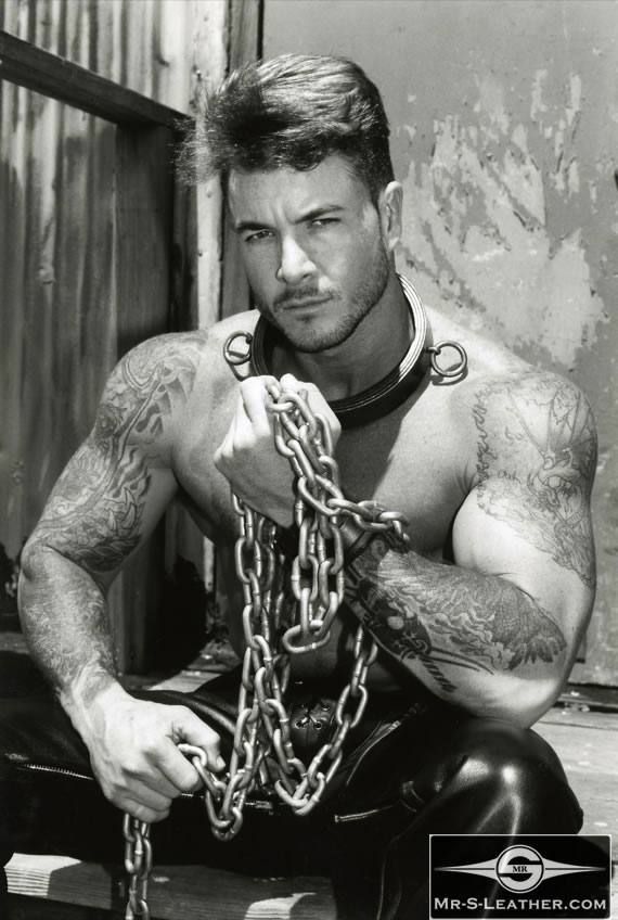 A shirtless, tattooed muscular man poses with heavy metal chains and a metal collar while wearing leather pants.