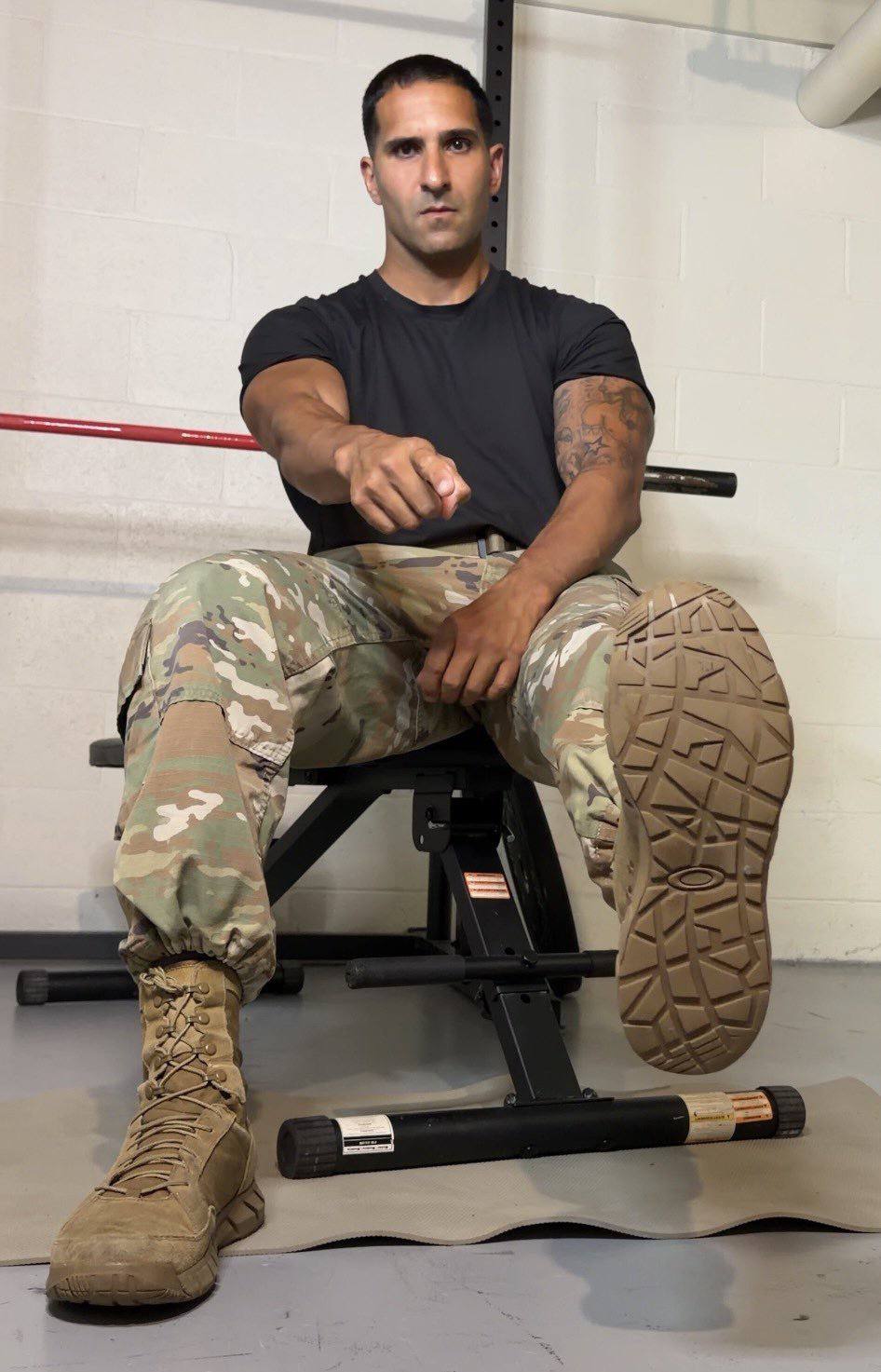 A muscular man in a black t-shirt and camouflage trousers sits on a gym bench, pointing at the camera while lifting the sole of his combat boot.