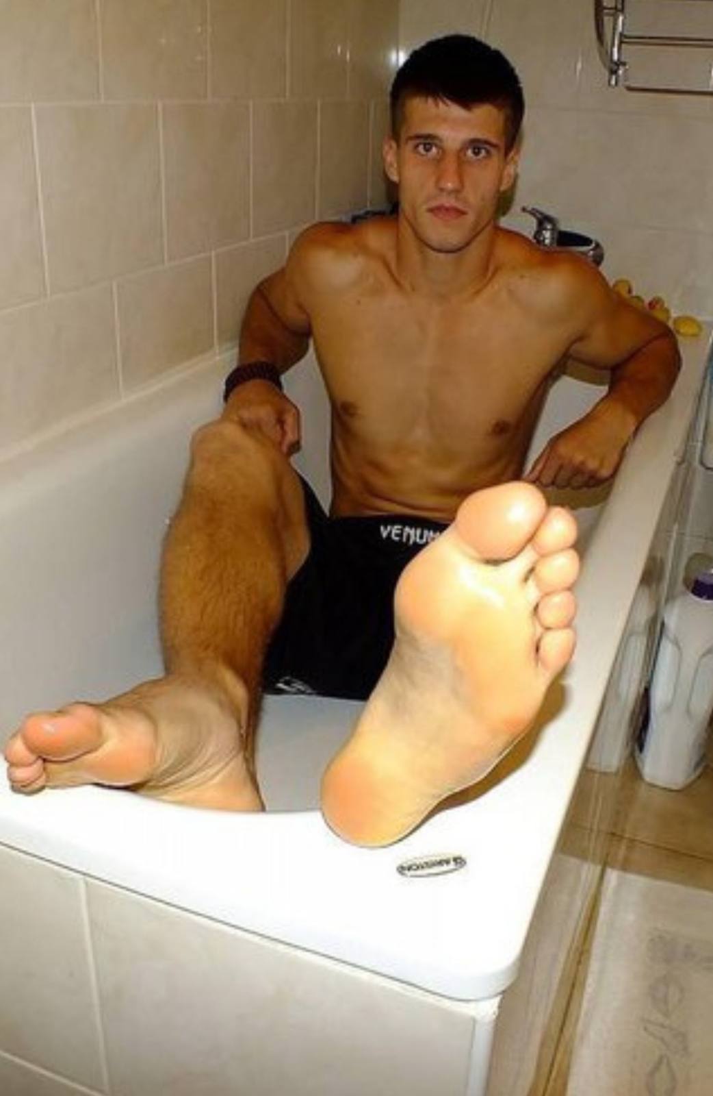 A shirtless muscular twunk sits in a bathtub wearing black athletic shorts, prominently displaying his bare feet and soles toward the camera.