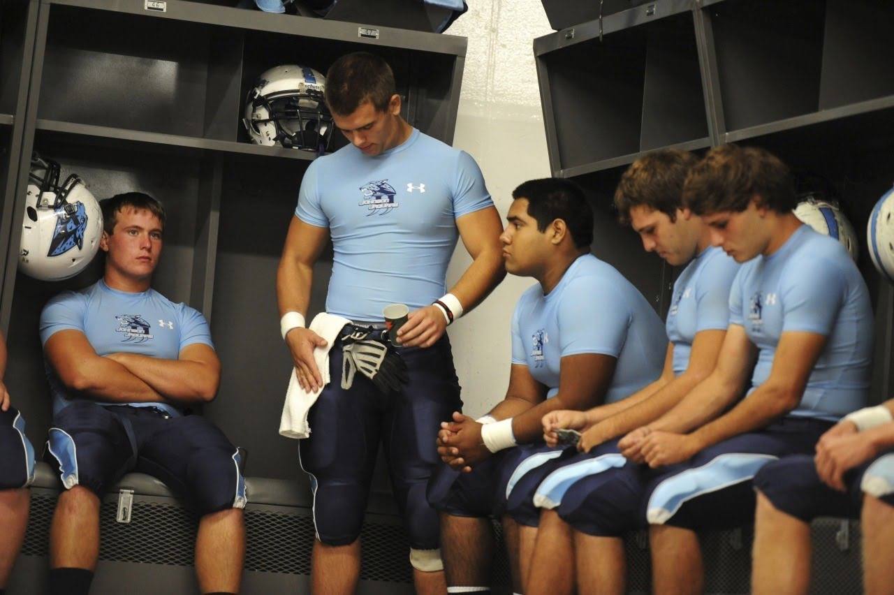 A group of athletic young football players wearing sky blue compression tops and padded football pants inside a locker room.