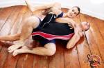 Two athletic men wearing wrestling singlets are grappling on a wooden floor in an indoor setting.