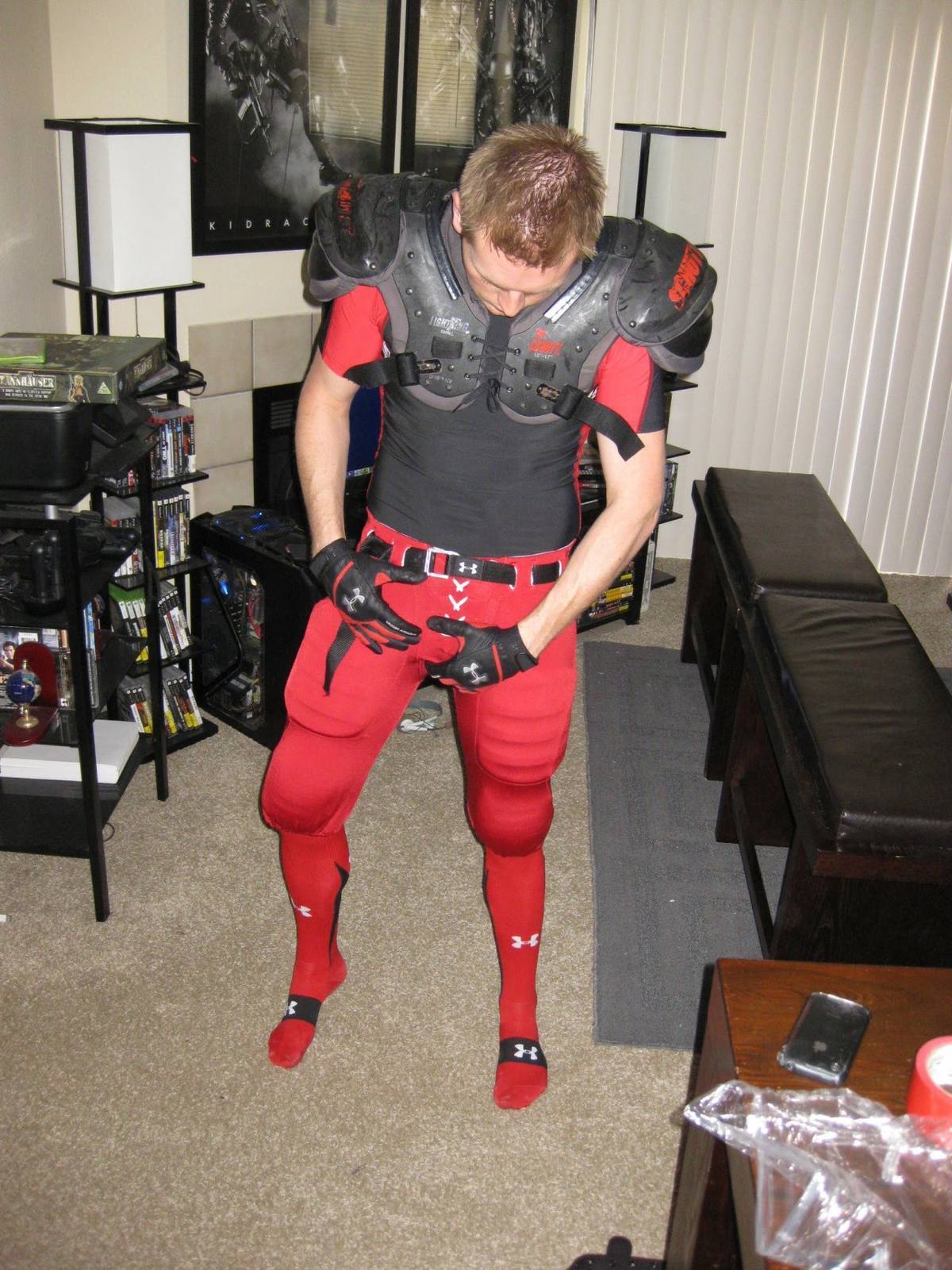 A twunk dressed in a full red and black American football kit with shoulder pads poses solo in a living room.