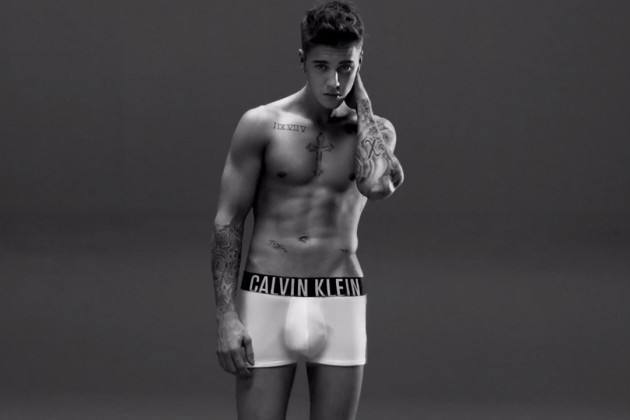 A young man poses shirtless in white boxer briefs with a prominent waistband.