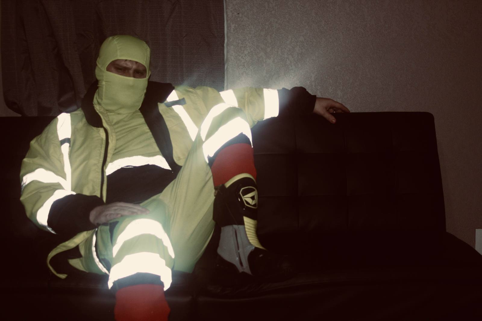 A man sits on a dark couch wearing a full high-visibility construction uniform and a matching balaclava that hides his face.