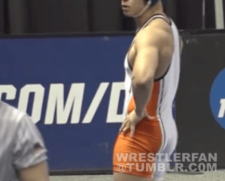 A male wrestler with an athletic body type stands wearing an orange and white wrestling singlet.