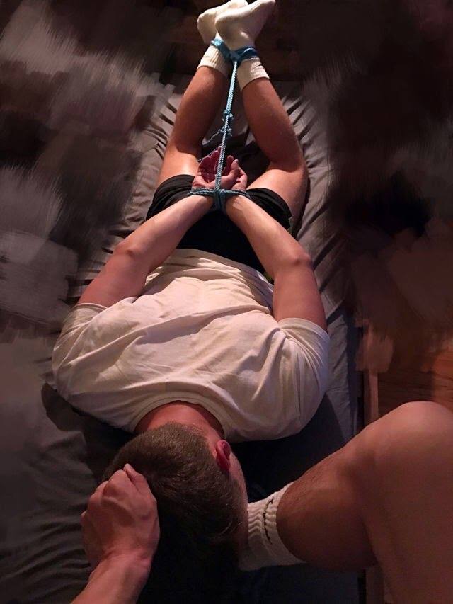 A man in gym shorts and white tube socks is hogtied with blue rope on a bed while a dominant partner holds his head.