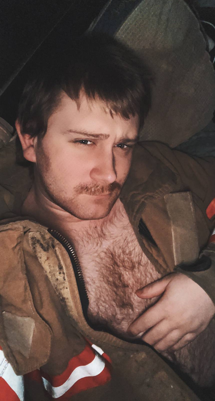 A masculine man with a hairy chest and mustache poses while wearing an unzipped tan firefighter jacket.