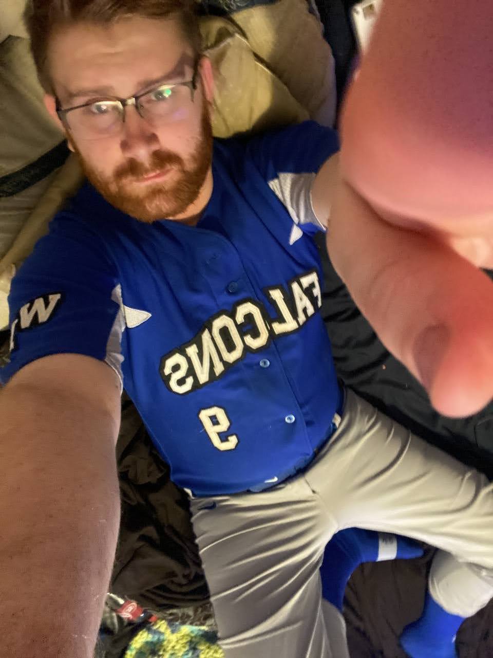 A bearded man in glasses wearing a blue Falcons baseball jersey and grey pants lies on a bed.